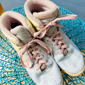 May include: A pair of light blue high-top trainers with pink laces and a yellow and white interior. The trainers are sitting on a blue and white patterned surface.
