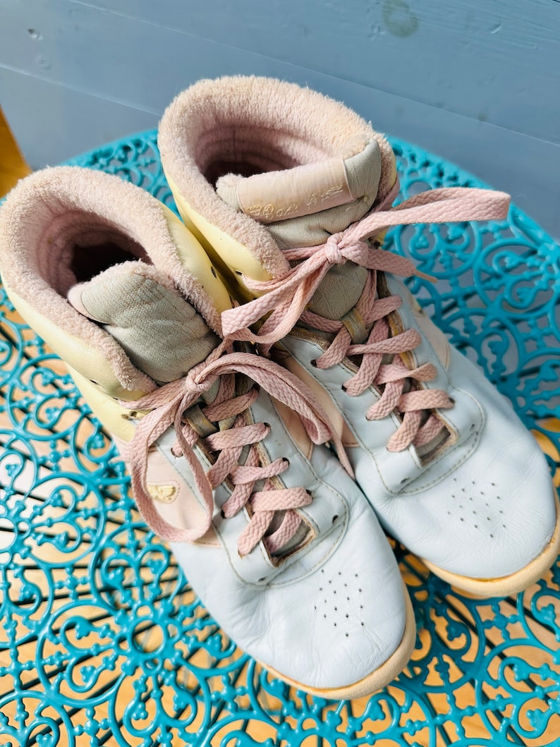 May include: A pair of light blue high-top trainers with pink laces and a yellow and white interior. The trainers are sitting on a blue and white patterned surface.