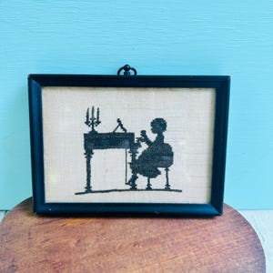 May include: A rectangular black frame containing a cross-stitch embroidery on light beige fabric. The embroidery depicts a black silhouette of a person sitting at a desk with a candelabra, rendered in fine black thread against the neutral background.