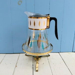 Pode incluir: Uma jarra de vidro vintage com uma faixa branca com desenhos de folhas douradas. A jarra tem uma alça preta e está colocada em um suporte de metal dourado. Folhas douradas e turquesas decoram o vidro. Uma peça clássica para servir bebidas.