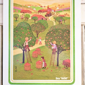 Puede incluir: Una ilustraci&oacute;n vintage de un huerto de manzanos rural con personas recogiendo manzanas de los &aacute;rboles. La ilustraci&oacute;n presenta un granero rojo, un camino sinuoso y un campo verde con manzanas rojas esparcidas por el suelo. El texto "Sun Valley" est&aacute; impreso en la esquina inferior derecha.