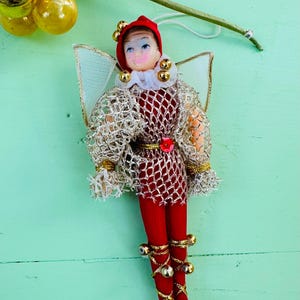 May include: A vintage Christmas bauble depicting a jester in a red and gold costume, white collar, and gold bells on its feet. The bauble has a gold mesh overlay and a white mesh wing.