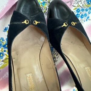 Puede incluir: Un par de zapatos de tac&oacute;n de ante y cuero negro con detalles dorados. Los zapatos tienen punta puntiaguda y tac&oacute;n bajo. La plantilla est&aacute; etiquetada con "Gucci Made in Italy".