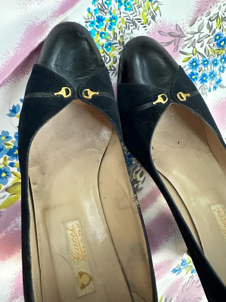 Puede incluir: Un par de zapatos de tac&oacute;n de ante y cuero negro con detalles dorados. Los zapatos tienen punta puntiaguda y tac&oacute;n bajo. La plantilla est&aacute; etiquetada con "Gucci Made in Italy".