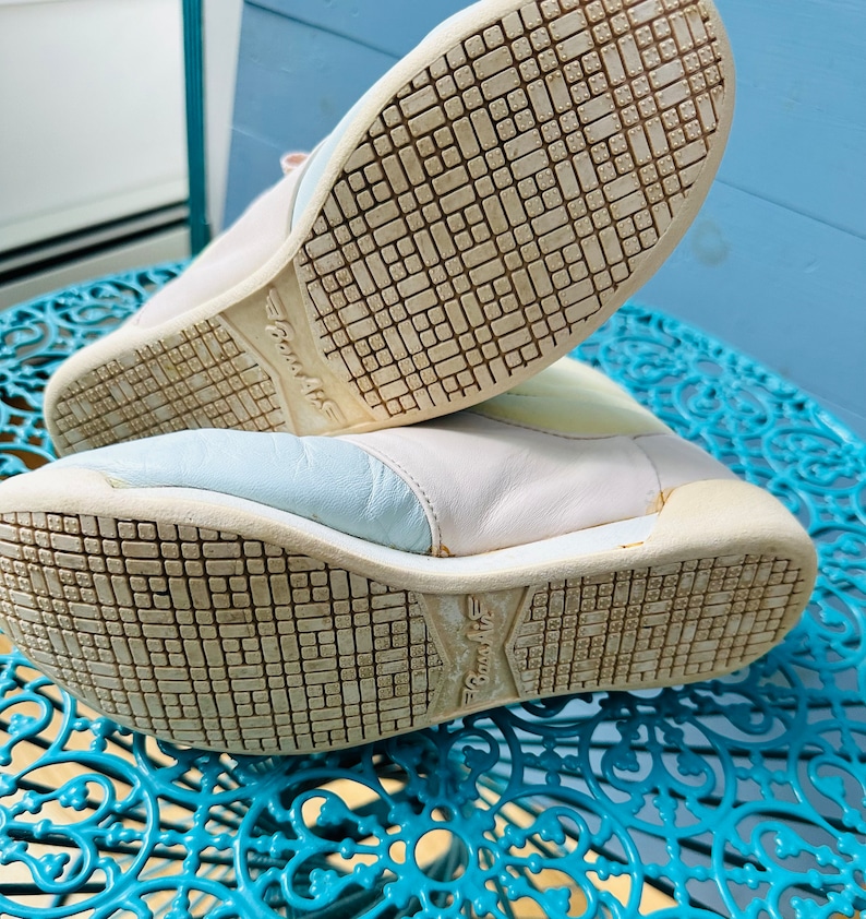 May include: A pair of white leather trainers with blue and yellow accents. The trainers have a textured sole with the word "Bounce Air" printed on the bottom.