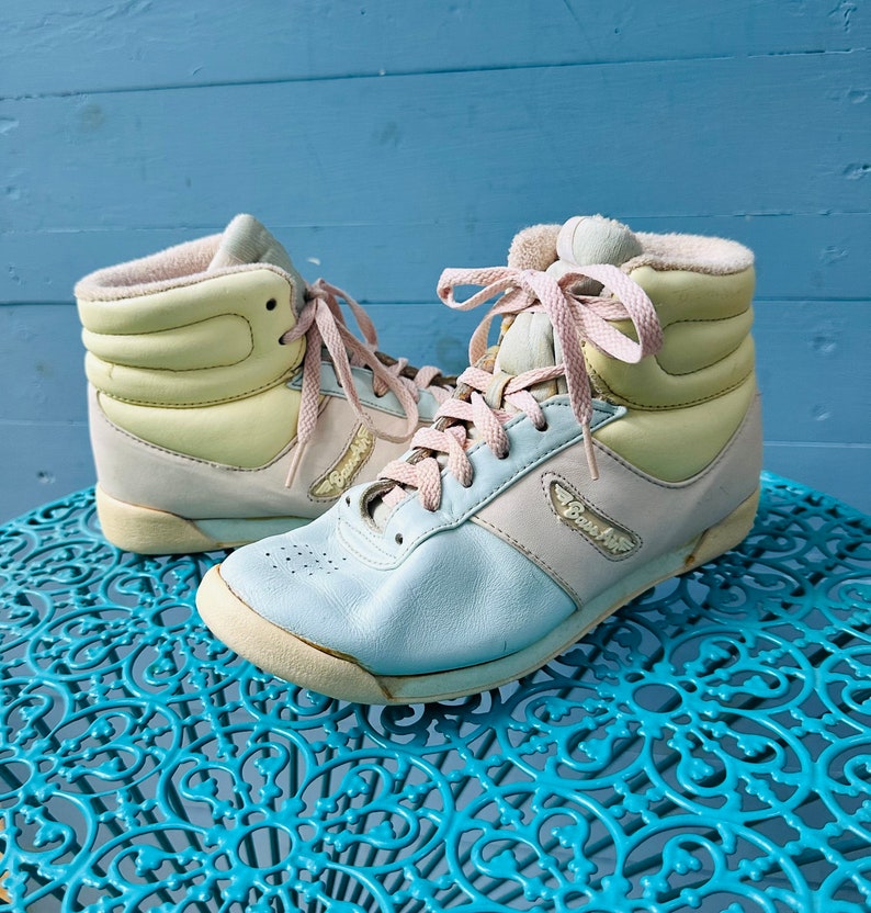 May include: A pair of high-top sneakers in pastel colors. The sneakers are light blue, pink, and yellow. The laces are pink. The sneakers are on a blue and white patterned surface.