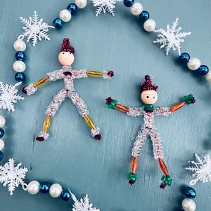 Op de afbeelding: Twee kralen-sneeuwpop-versieringen met rode mutsen en witte kralen. De versieringen zijn omringd door een witte parel en blauwe kralenkrans met witte sneeuwvlokaccenten.