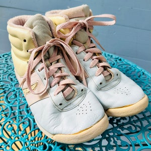 May include: A pair of pastel-coloured high-top trainers with pink laces. The shoes are made of leather and have a white sole.