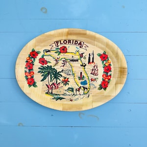 May include: An oval bamboo serving tray featuring a map of Florida with the text FLORIDA at the top. The map includes illustrations of palm trees, flamingos, hibiscus flowers, oranges, and various landmarks, all set against a light wood grain background.