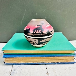 May include: A small, rounded ceramic vase featuring a hand-painted scene of a rider on horseback against a pink and grey sunset sky. The lower portion of the vase displays black geometric patterns on a tan background, creating a distinct contrast.