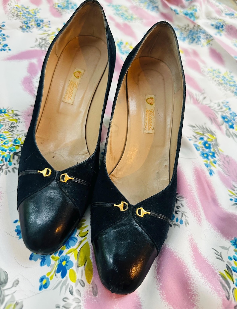 Puede incluir: Un par de zapatos de tac&oacute;n de ante y cuero negro con detalles dorados. Los zapatos tienen una punta redonda y una peque&ntilde;a hebilla dorada en la parte delantera. Los zapatos est&aacute;n sobre una tela de estampado floral blanco y rosa.