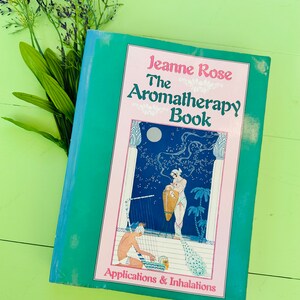 May include: A teal green book titled "Jeanne Rose The Aromatherapy Book" with a pink and white illustration of a woman holding a vase and a man playing a harp. The book's subtitle is "Applications & Inhalations".