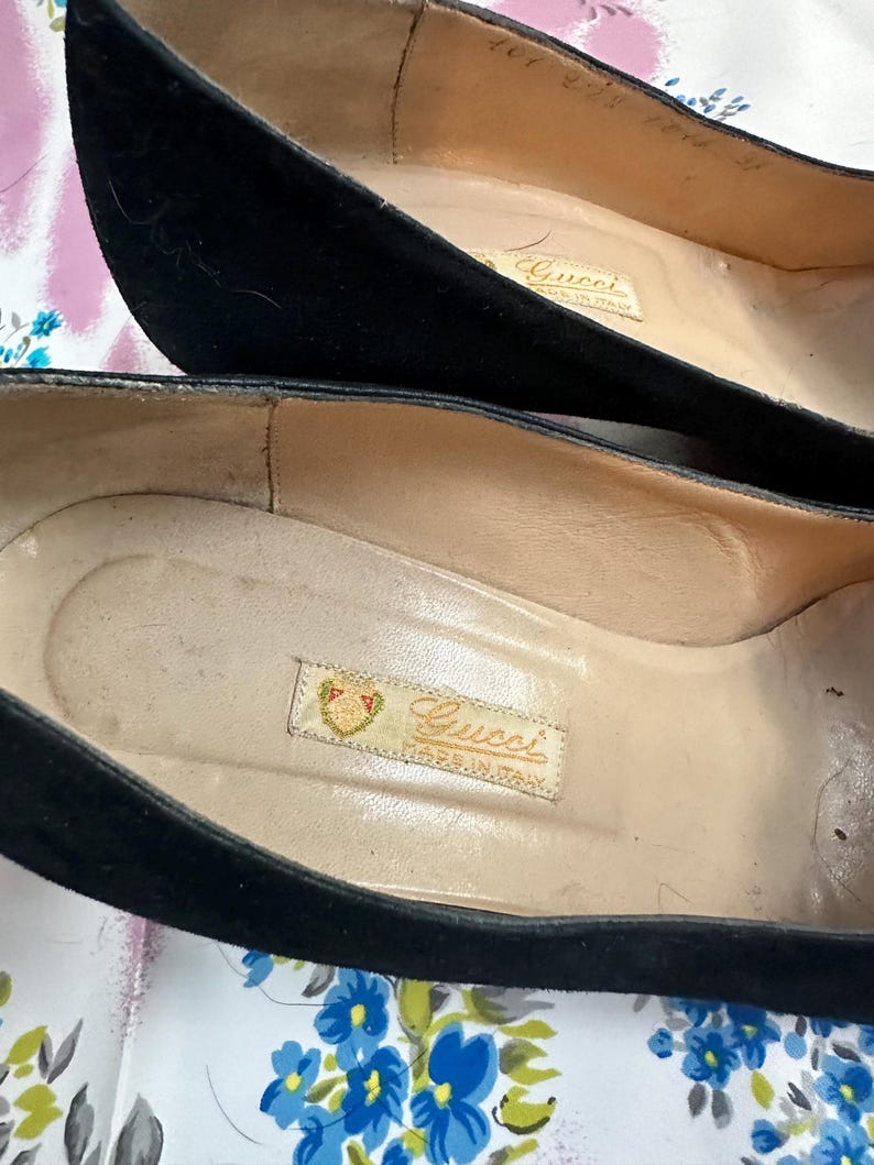 Puede incluir: Un par de zapatos de ante negro con una etiqueta de Gucci en el interior. La etiqueta dice "Gucci Hecho en Italia".