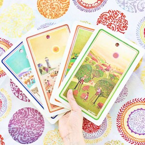Puede incluir: Cuatro tarjetas ilustradas coloridas con un borde verde, azul, amarillo y naranja. Las tarjetas presentan una variedad de escenas, incluyendo un faro, un pueblo y un campo de manzanos. Las tarjetas est&aacute;n dispuestas en forma de abanico y se sostienen en una mano.