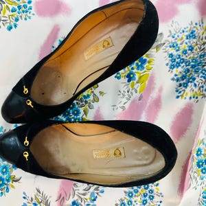 Puede incluir: Un par de zapatos de tac&oacute;n de ante negro con detalles dorados. Los zapatos tienen punta puntiaguda y cierre trasero. Los zapatos est&aacute;n sobre un fondo floral blanco y rosa.