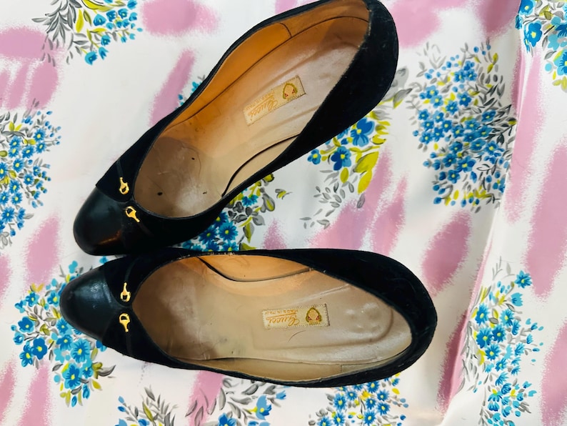 Puede incluir: Un par de zapatos de tac&oacute;n de ante negro con detalles dorados. Los zapatos tienen punta puntiaguda y cierre trasero. Los zapatos est&aacute;n sobre un fondo floral blanco y rosa.