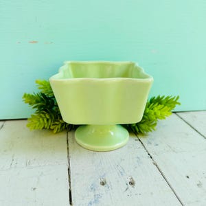 May include: A light green pedestal bowl with a square, scalloped rim and a smooth, glossy finish. The bowl features a central pedestal base and is crafted from a solid, opaque material, presenting a uniform color throughout its entire structure.