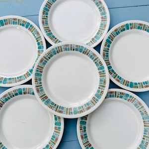 Puede incluir: Seis platos de cerámica blancos con un diseño de borde geométrico en turquesa, marrón y verde. Los platos están dispuestos sobre una superficie azul claro, mostrando una estética vintage. Los platos son adecuados para servir comidas.