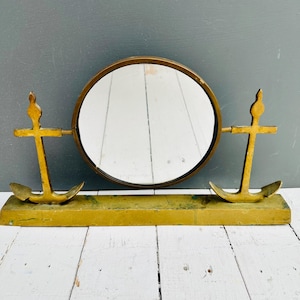 May include: A vintage brass vanity mirror with a nautical theme. The round mirror is framed in brass and flanked by two brass anchor designs. The mirror sits on a rectangular base, creating a decorative and functional piece.