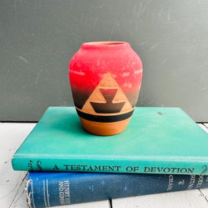 Peut inclure: Petit vase en terre cuite avec un motif géométrique rouge et noir. Le vase présente un motif triangulaire avec une bande noire et est posé sur un fond de livres anciens.