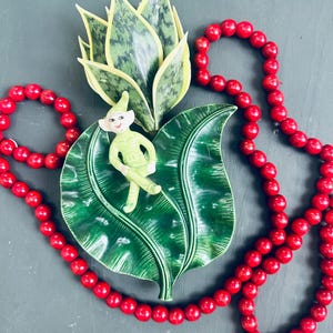 May include: A green ceramic leaf dish with a green elf figurine sitting on top. The dish is surrounded by a strand of red beads.
