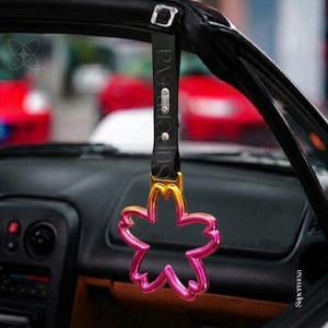 Könnte beinhalten: Ein Auto-Haltegriff mit einem kirschblütenförmigen Ring in einem Farbverlauf von Gelb zu Pink. Der Ring ist an einem schwarzen Riemen mit einer silbernen Metallschnalle und einer kleinen silbernen Platte befestigt, die mit Supernova graviert ist.