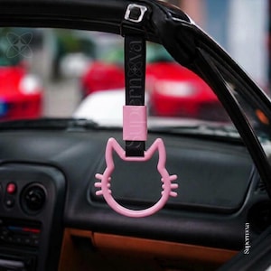 May include: A pink cat-shaped car handle with a black strap featuring the text Supernova. The handle is suspended from the interior roof of a vehicle, showcasing a cat silhouette with ears and whiskers, attached by a rectangular pink connector.