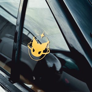 Peut inclure: Un autocollant de fenêtre de voiture jaune et noir avec un graphisme stylisé d'une tête de créature avec une étoile au sommet.