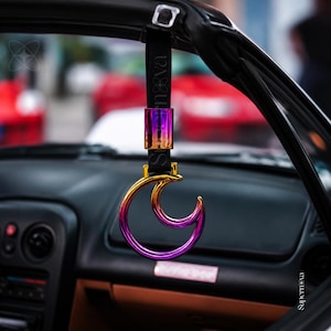 May include: A crescent moon-shaped car handle with an iridescent, multi-colored metallic finish. The handle is attached to a black strap featuring the text Supernova and a small logo. The strap is secured to the interior frame of a vehicle.
