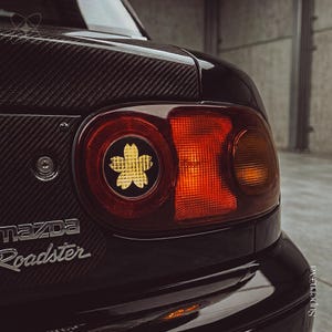 Peut inclure: Une section arrière de voiture Mazda Roadster avec un coffre en fibre de carbone et un feu arrière rouge circulaire. Un autocollant de fleur de cerisier doré est centré dans le boîtier du feu circulaire, avec l'emblème Mazda Roadster visible.