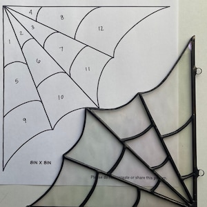 Pode incluir: Um desenho a preto e branco de uma teia de aranha com seções numeradas. O desenho está numa folha de papel branca com o texto "Spider Web" e "Pattern Created By GlassHalfEmptied" no topo. O desenho mede 8 polegadas por 8 polegadas. O texto "Hobby License" está num círculo no canto superior direito do papel. Uma teia de aranha de vitral preto é mostrada no canto inferior direito da imagem.