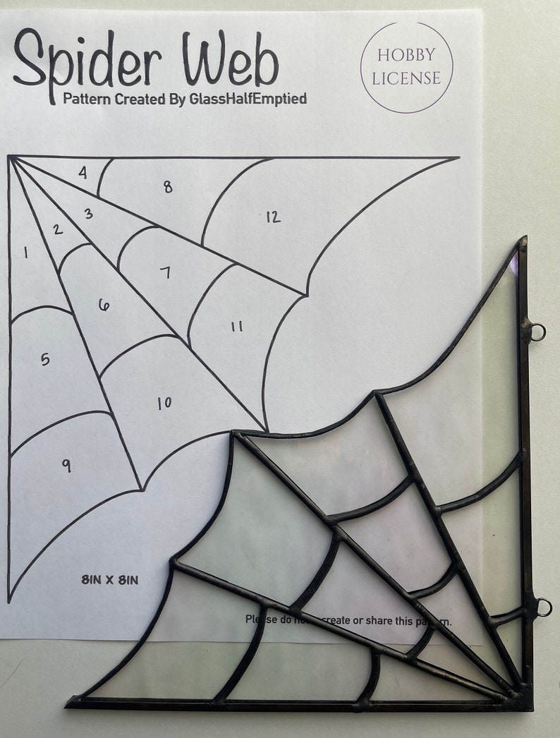 May include: A black and white line drawing of a spider web with numbered sections. The drawing is on a white sheet of paper with the text "Spider Web" and "Pattern Created By GlassHalfEmptied" at the top. The drawing is 8 inches by 8 inches. The text "Hobby License" is in a circle at the top right corner of the paper. A black stained glass spider web is shown in the lower right corner of the image.