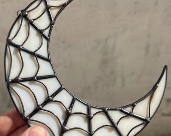 May include: A stained glass crescent moon with a spiderweb design. The moon is white with black lines and a metal frame. The decorative piece is held by a hand.