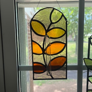 May include: A stained glass window hanging with a clear glass tube for a flower vase. The stained glass features a leafy design in shades of yellow, orange, and brown.