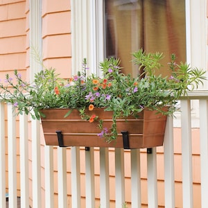 May include: Two copper planter boxes with black metal brackets are mounted on a white wooden railing. The planter boxes are filled with a variety of colourful flowers and greenery.