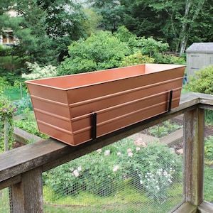 May include: A copper-colored metal planter box with a wood railing and a wire mesh fence in the background. The planter box has a rectangular shape and is mounted to the railing with black metal brackets.