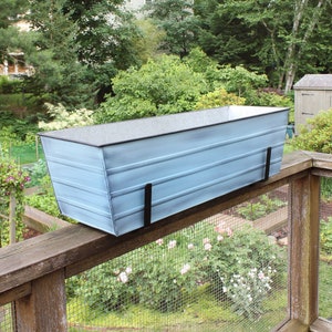 May include: A light blue metal planter box with black metal brackets, mounted on a wooden railing. The planter is hanging over a green garden with a wire fence.