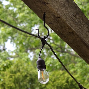 May include: A black metal hook attached to a wooden beam with a black wire running to a light bulb. The light bulb is clear glass with a black rubber coating and is lit.