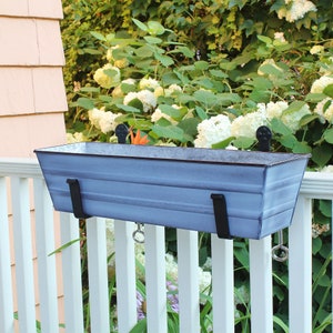 Peut inclure: Un pot de fleurs en métal bleu clair avec un design ondulé est suspendu à une balustrade blanche. Le pot a deux supports en métal noir et deux anneaux en métal sur les côtés.