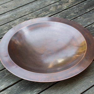 May include: A large, round copper fire pit with a raised rim. The fire pit is empty and has a slightly weathered finish.