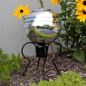 May include: A reflective glass gazing ball with blue stripes, mounted on a black metal stand with three legs. The sphere reflects the surrounding environment, including yellow flowers and greenery. The garden ornament is placed in a garden bed.