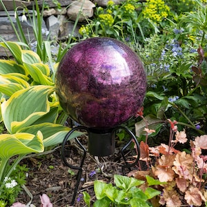 May include: A decorative, spherical garden gazing ball with a textured, iridescent purple finish. The ball sits atop a black, wrought-iron stand with three curved legs. The garden ornament is surrounded by lush green and colorful foliage.
