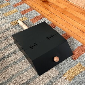 May include: Black metal dustpan with a wooden handle. The dustpan has two hinges and a wooden knob on the side.