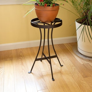May include: Black metal plant stand with a round top and three legs. A terracotta pot with red flowers sits on top of the stand.