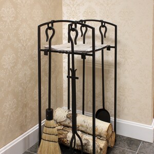 May include: Black metal fireplace tool holder with a broom, shovel, poker, and tongs. The holder has a shelf for storing logs and a newspaper. The holder is standing on a tiled floor.