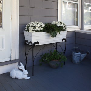 May include: White planter box with white flowers and green plants on a black metal stand with a white rabbit figurine on a gray porch.