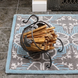 May include: A black metal log holder with a wire mesh basket filled with wooden logs. The holder is on a gray and blue patterned rug.
