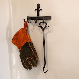 May include: A black metal fireplace tool set hanging on a wall. The set includes a poker, a glove, and a hanger with hooks. The glove is orange and black. The poker has a decorative heart-shaped handle.