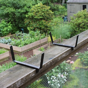 May include: Two black metal plant brackets mounted on a wooden fence rail. The brackets are designed to hold a plant pot or planter.