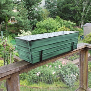 May include: A green metal planter box with a galvanised finish is mounted on a wooden railing. The planter is hanging over a garden with green plants and flowers.